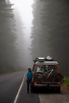 a person standing next to a van with bikes on the back in the foggy forest