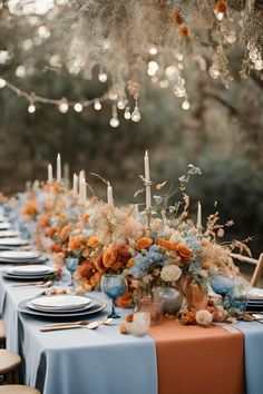 a long table is set with blue and orange flowers, candles, and place settings