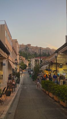 people are walking down an empty street in front of some buildings and hills on the hill behind them