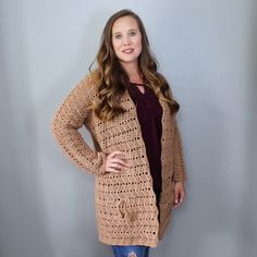 a woman standing in front of a gray wall wearing a brown cardigan and jeans