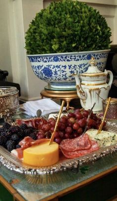 a tray with cheese, meats and grapes on top of a glass table in front of a potted plant