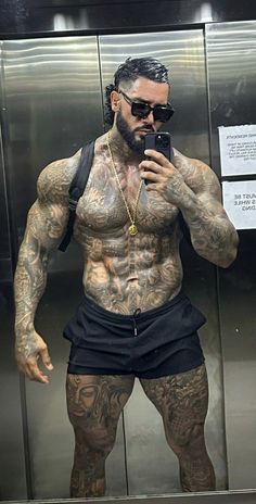 a man with tattoos taking a selfie in front of a mirrored wall and holding a cell phone