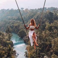 a woman is swinging from a rope in the air over a river and jungle area