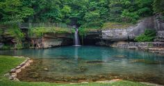 there is a small waterfall in the middle of this lake with green trees around it