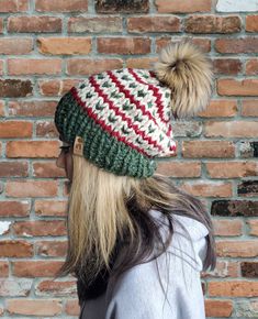 a woman with long hair wearing a green and white knitted hat, standing in front of a brick wall