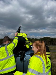 two people in yellow jackets standing next to a car with their hands on the steering wheel