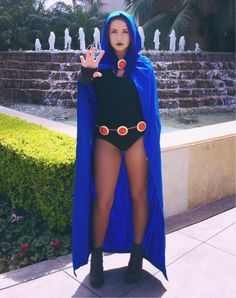 a woman dressed in a costume standing on the sidewalk with her hand up and wearing a blue cape