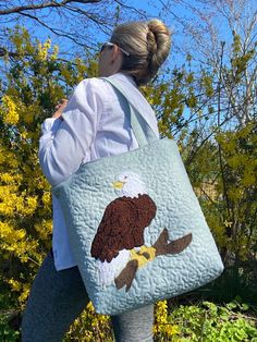 "This handmade bag has a beautiful bold Bald Eagle perched on a branch. This bag was made in mind for all of those eagle nest followers. So many famous nest cams are watched today. I have followed several nest over the years and have been inspired to make these bags. This bag makes a great gift and will last for many years.  The bag is approximately 13\"x 14\". It is extremely durable and lined. It has 3 inside pockets. It also has a magnetic clasp that opens and closes easily. The bag has nice long padded handles approximately 27 \" long. The front of the bag is quilted. Each bag I make is one of a kind because it is done freehand by me. The bags are also hand pieced and stitched in our smoke free home. Free shipping included with this purchase." Quilted Rectangular Bag For Gifts, Quilted Rectangular Bag For Gift, Quilted Rectangular Bag As Gift, Rectangular Quilted Bag For Gifts, Rectangular Quilted Bag As Gift, Quilted Pouch Bag As Gift, Quilted Pouch Bag For Gift, Eagle Quilt, Eagle Nest