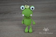 a crocheted frog keychain hanging from a chain on a wooden surface