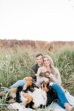 a man and woman are sitting in the grass with two dogs, one is holding a baby