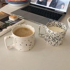 two coffee mugs sitting next to each other on a desk with a laptop in the background