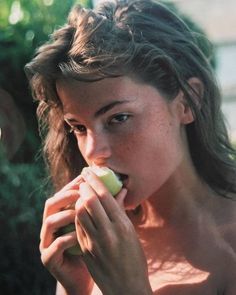 a woman is eating an apple outside in the sun with her shirt off and one hand on her face