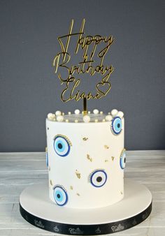 a birthday cake with an evil eye design and gold happy birthday sign on top, sitting on a white plate