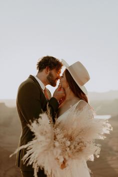 a bride and groom kissing in the desert with their hat on his head, wearing white feathers