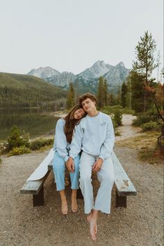 two people sitting on a bench in front of a body of water with mountains in the background