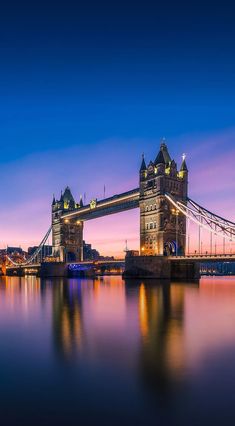 the tower bridge is lit up at night with its lights on and reflecting in the water