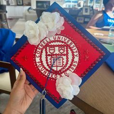 a person holding up a red and white graduation cap with flowers on it in front of a table