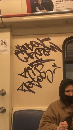 a man wearing a hoodie sitting on a train with graffiti written on the wall behind him