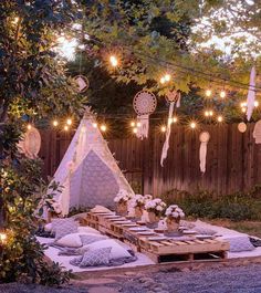 an outdoor picnic with lights strung from the trees and pillows on the ground, in front of a wooden fence