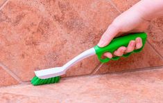 a hand holding a green and white brush on top of a tile floor next to a tiled wall