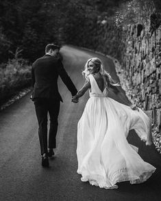 black and white photo of bride and groom holding hands walking down the road in front of a stone wall