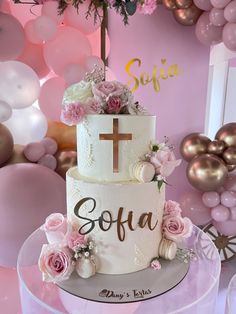 a white cake with pink flowers and a cross on top is sitting on a table in front of balloons