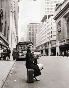 black and white photograph of woman sitting on suitcase in street with city bus behind her