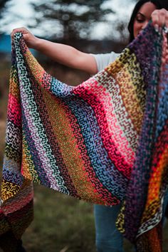 a woman holding up a multicolored crocheted shawl in her hands