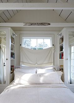 a bed with white linens and pillows in a bedroom next to two large windows