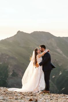 a bride and groom kissing on the top of a mountain in front of mountains with their arms around each other