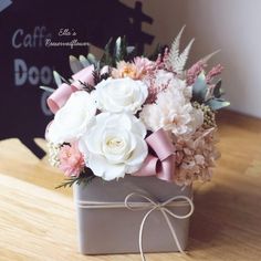 a bouquet of white and pink flowers in a gray box on a wooden table with a sign behind it