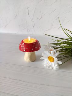 a mushroom shaped candle holder next to a daisy on a white surface with a plant in the background