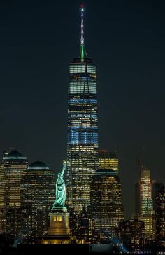 the statue of liberty is lit up at night in new york city, with skyscrapers behind it