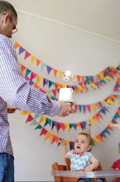 a man holding a small cake in front of a baby sitting at a table with flags on the wall behind him