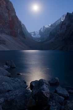 the moon is setting over a mountain lake with rocks and boulders in front of it