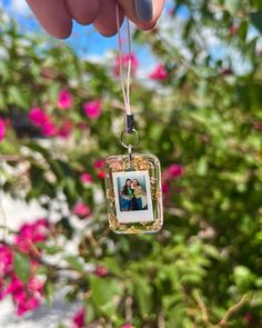 a hand holding a small square photo ornament in front of some pink flowers