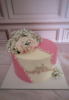 a white and pink cake with flowers on top sitting on a table next to a wall