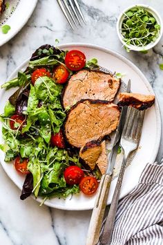 a white plate topped with meat and salad on top of a marble table next to silverware