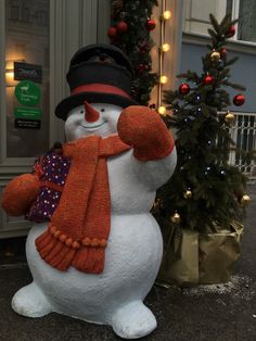 a large snowman with a hat and scarf standing next to a christmas tree in front of a building