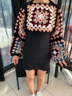 a woman standing in front of a window wearing a crocheted sweater and sneakers