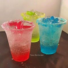 three different colored drinks sitting on top of a black table with ice cubes in them