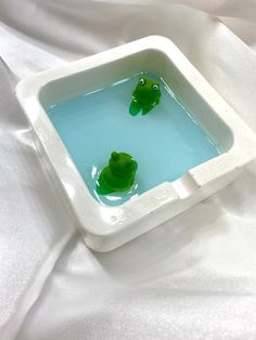 two little green frogs in a white square bowl filled with blue water on a white sheet