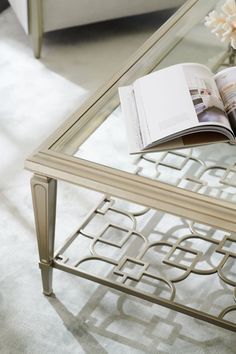 an open book is sitting on a glass coffee table with flowers in the corner and a white couch behind it