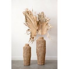 two tall vases with dried plants in them on a concrete floor next to a white wall