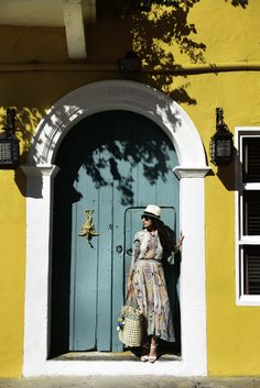 a woman standing in front of a blue door wearing a dress and hat with her hand on her hip