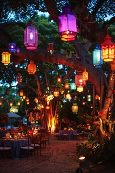 many lanterns are lit up in the trees at night time, with tables and chairs under them