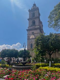 an old church tower towering over a garden filled with flowers and trees on a sunny day