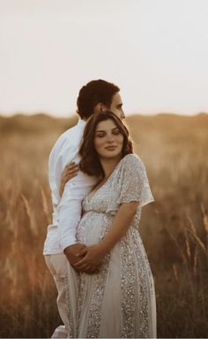 a pregnant woman standing next to a man in a field with tall grass at sunset