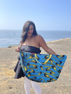 a woman carrying a blue and yellow bag on the beach by the ocean with her hands in her pockets