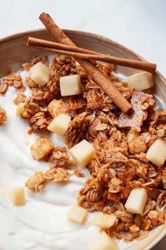 a bowl filled with granola and apples on top of a white countertop next to cinnamon sticks